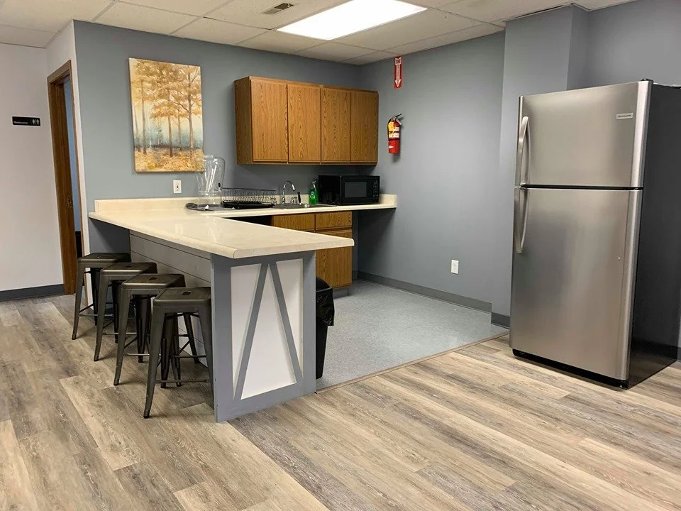 A small kitchenette with a gray wall, wooden cabinets, stainless steel refrigerator, microwave, a sink, and a countertop with four bar stools.