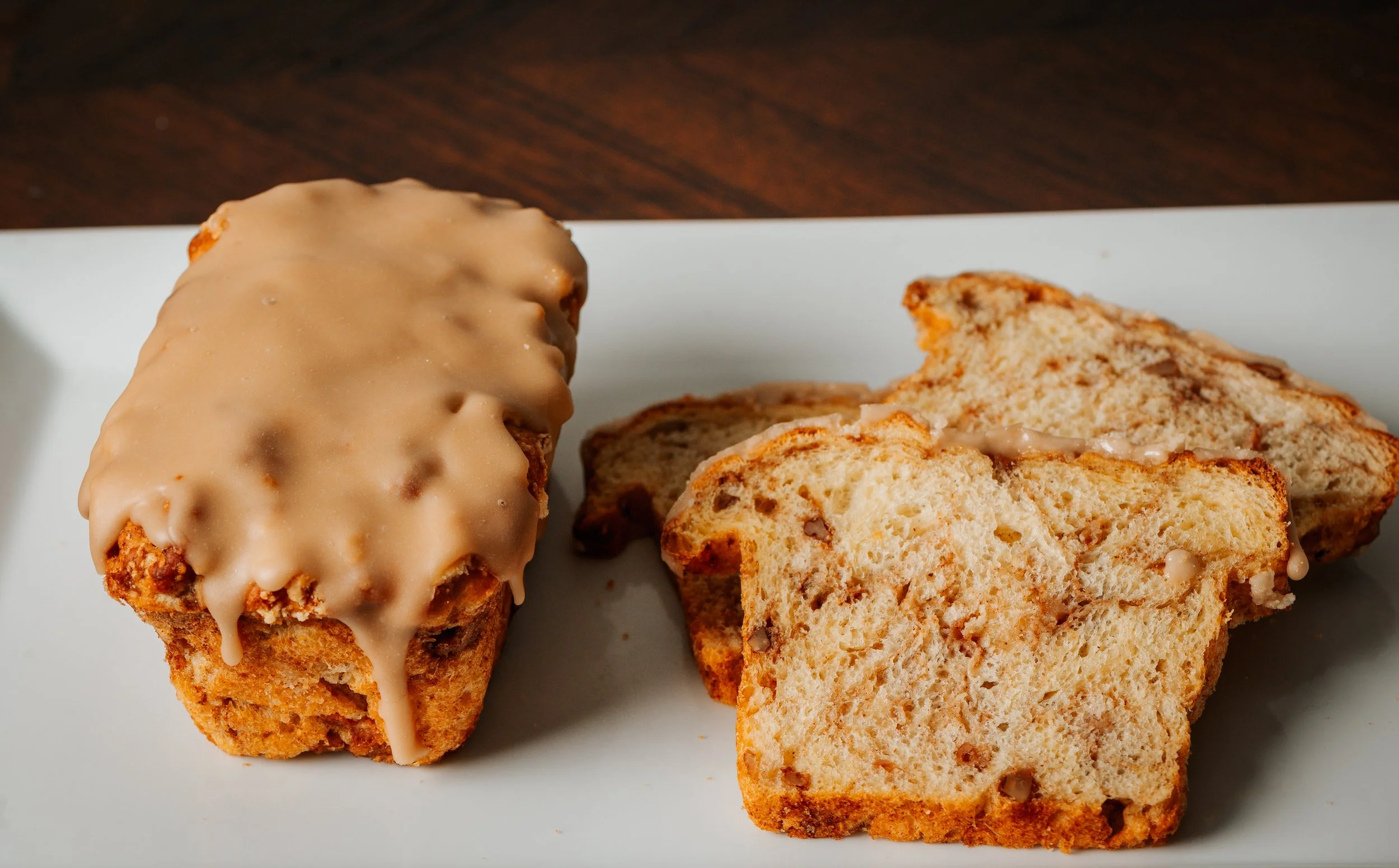 A loaf of Dutch Apple Bread is shown with a stack of slices next to it, on a white plate with a dark wooden surface in the background.