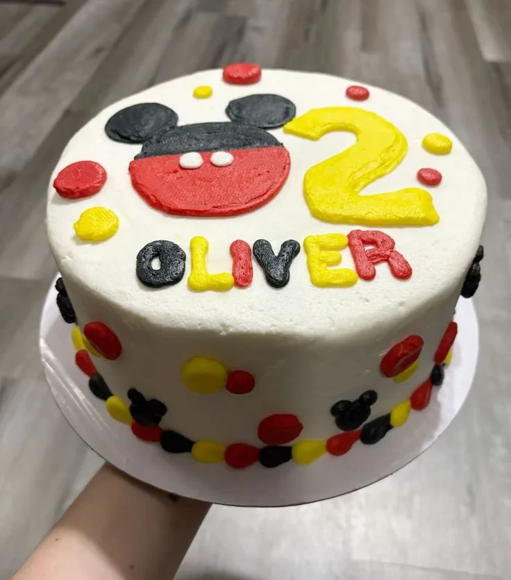 Cake decorated with a design resembling Mickey Mouse, the number 2, and the name Oliver, with red, yellow, and black polka dots around the sides.