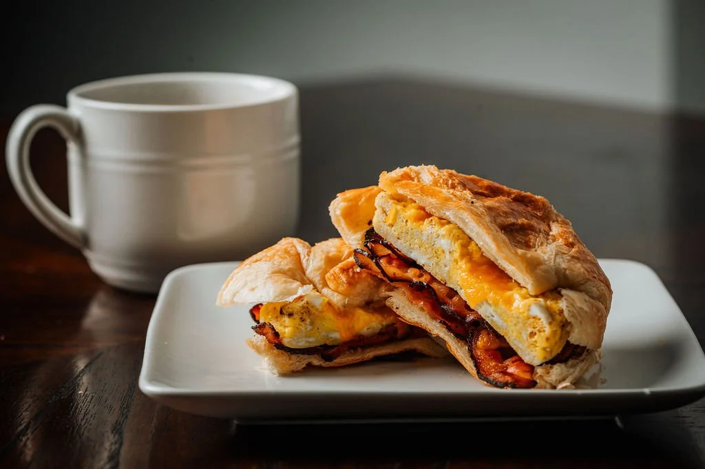 A breakfast sandwich with fried egg, bacon, and cheese on a croissant, served on a white plate, with a white mug of coffee in the background.