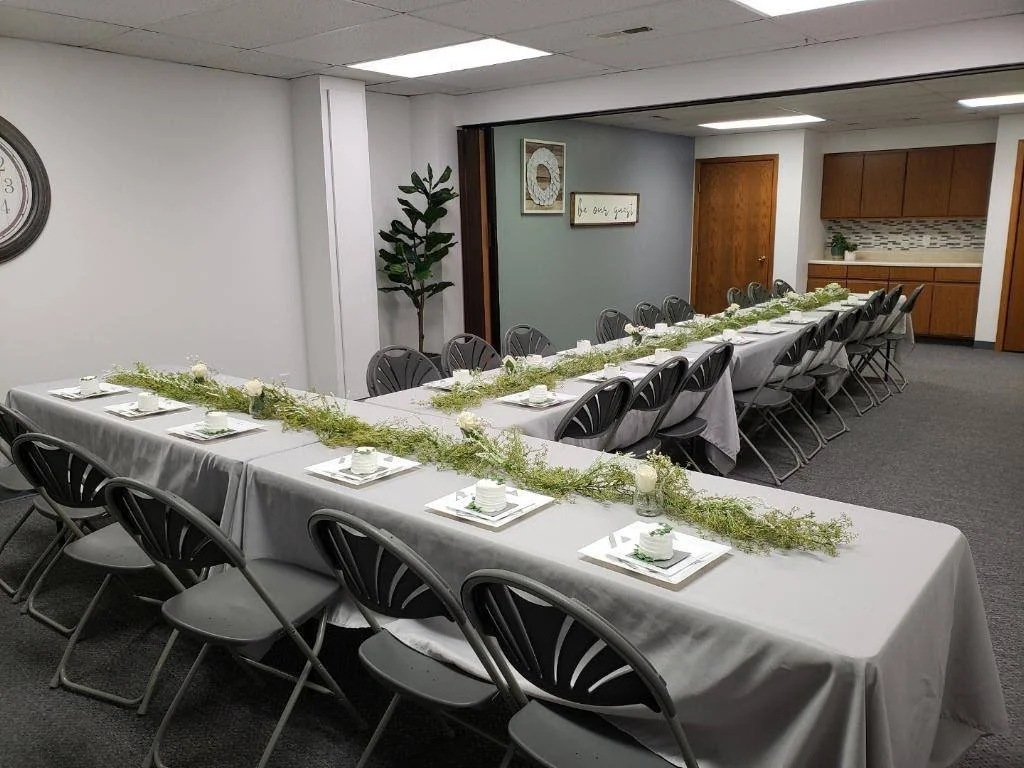 Long rectangular banquet table with white tablecloth and a green floral centerpiece, surrounded by black chairs, in a room with gray walls, wood accents, and decorative wall art.