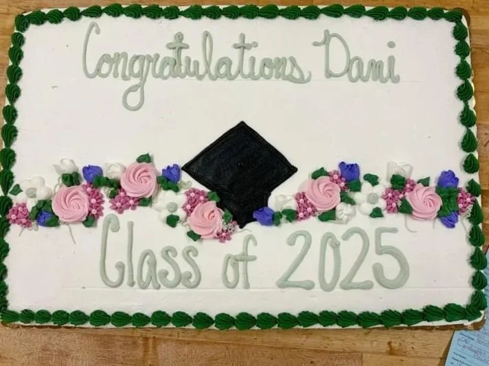 A graduation cake decorated with pink, purple, and white flowers, a black graduation cap, and green piping, with the message 'Congratulations Dani' and 'Class of 2025' written in icing.