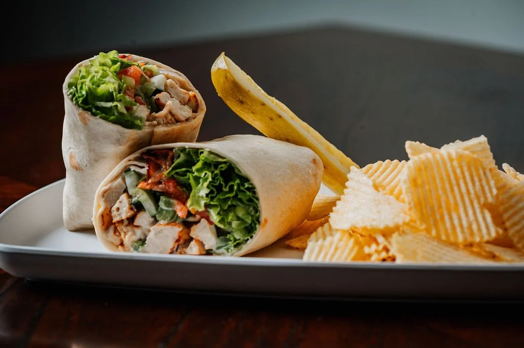 Close-up of a plate holding a chicken wrap cut in half, potato chips, and a pickle spear on a dark table.