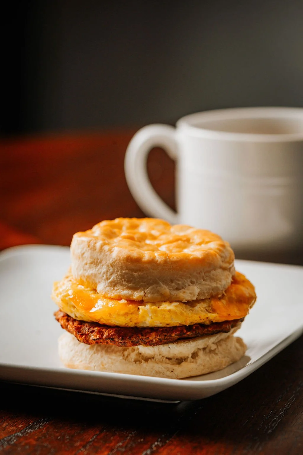Breakfast sandwich with egg, sausage patty, and biscuit on a white plate, with a white mug in the background.