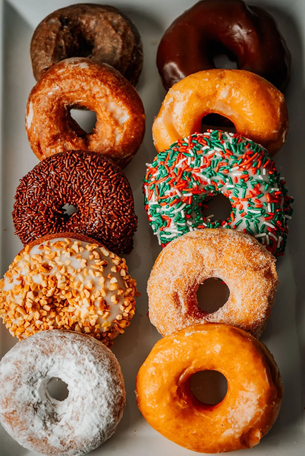 Assorted donuts with various toppings including chocolate sprinkles, chopped nuts, colorful sprinkles, and powdered sugar.