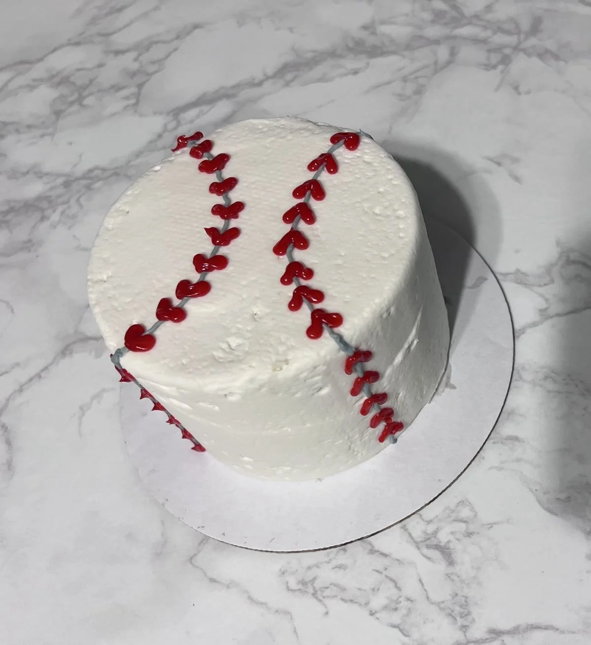 A white cake decorated in a baseball stitching pattern on top, sitting on a marble surface.