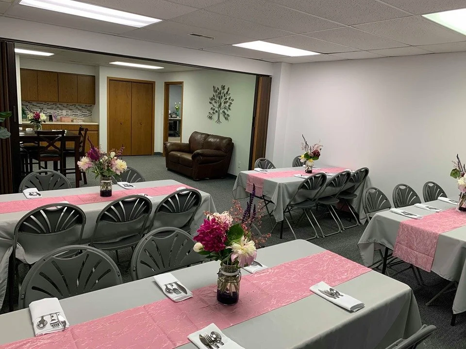 Room set up for a gathering with rectangular tables covered in white tablecloths and pink runners, each decorated with floral centerpieces in vases. There are gray plastic chairs around the tables, and the background shows a small kitchen area, a lea