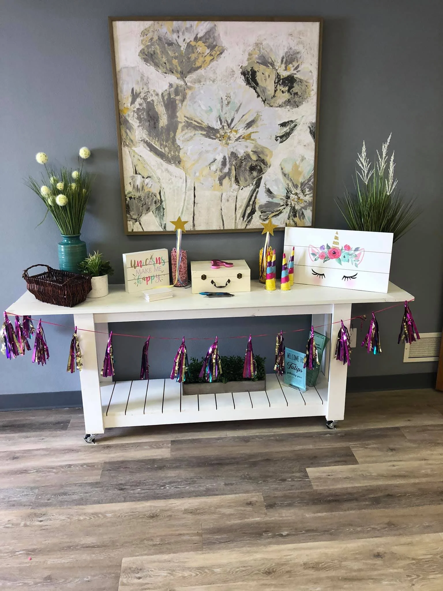 A white table decorated for a celebration with a purple and pink tinsel garland, a potted plant, a white box with smiley face, colorful party hats, a sign that says 'Welcome to Tulips', and two vases with decorative grass and flowers. Large floral pa