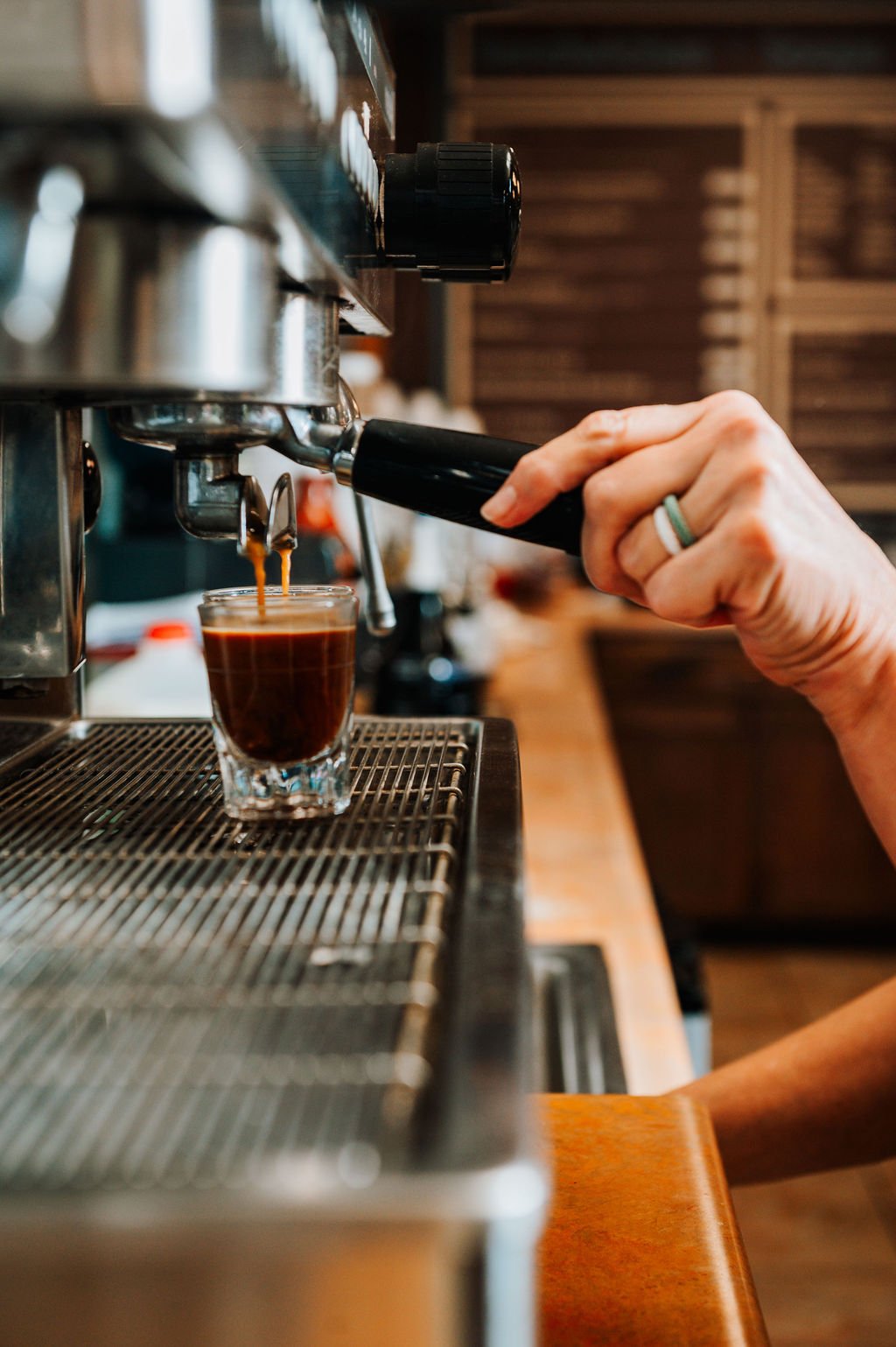 Close-up of a barista's hand pressing a lever on an espresso machine, brewing a shot of espresso into a small glass cup.