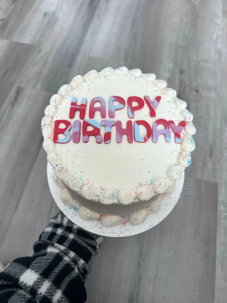 A birthday cake with white frosting, decorated with colorful sprinkles, and the words 'HAPPY BIRTHDAY' written in red and purple letters on top, being held by a person wearing a black and white checkered shirt, against a gray wooden floor background.