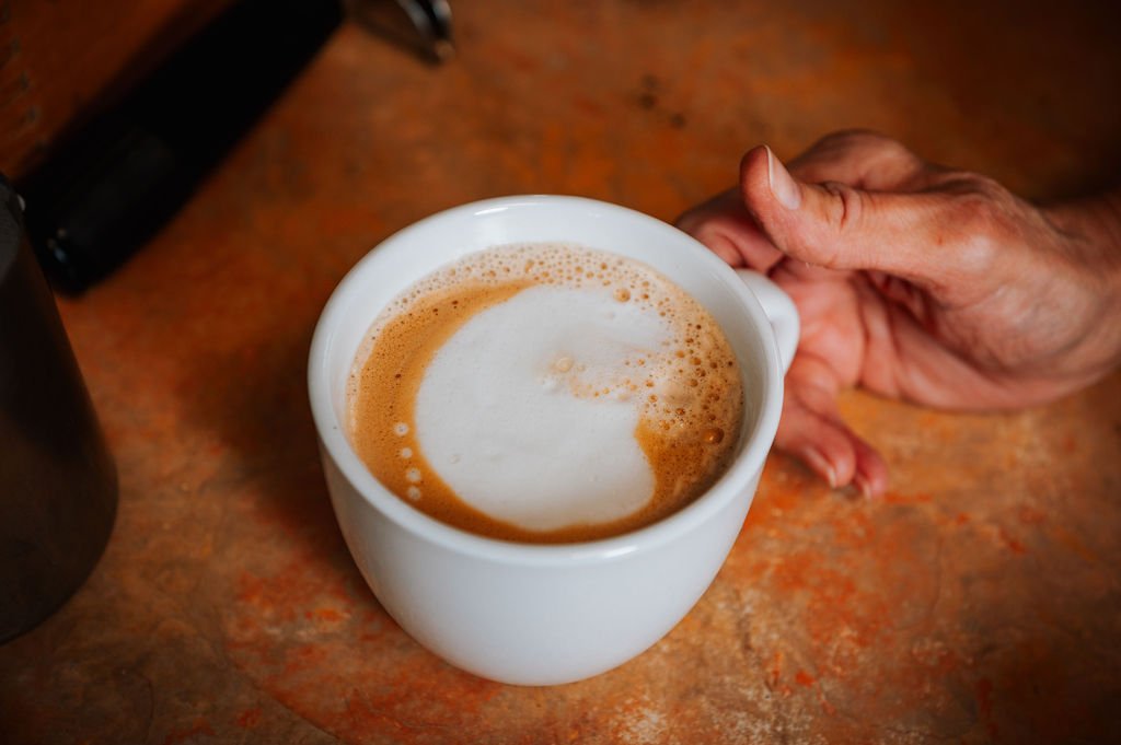 A person holding a white mug of coffee with foam on top, resting on a wooden table.
