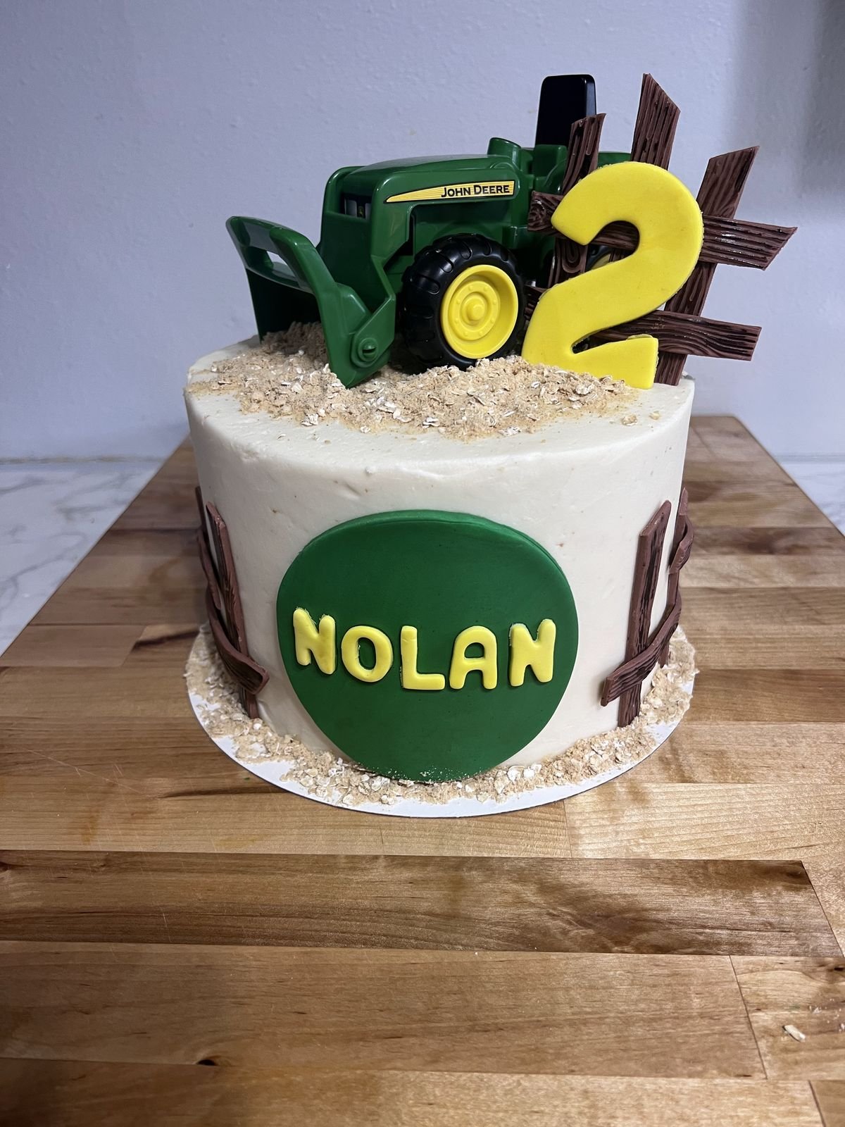 A cake with a tractor and nature theme, featuring a green John Deere tractor, a yellow number two, and a green circle with the name 'NOLAN' in yellow letters, decorated with brown fence-like accents and a textured ground topping.