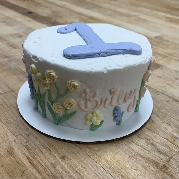 A round white birthday cake decorated with pastel-colored flowers and green leaves, with a blue 1 on top, on a wooden surface.