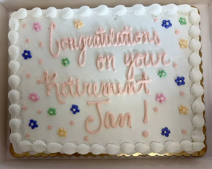A rectangular white birthday cake with white icing border, decorated with colorful small flower-shaped candies, and pink icing text that reads 'Congratulations on your retirement, Jan!'