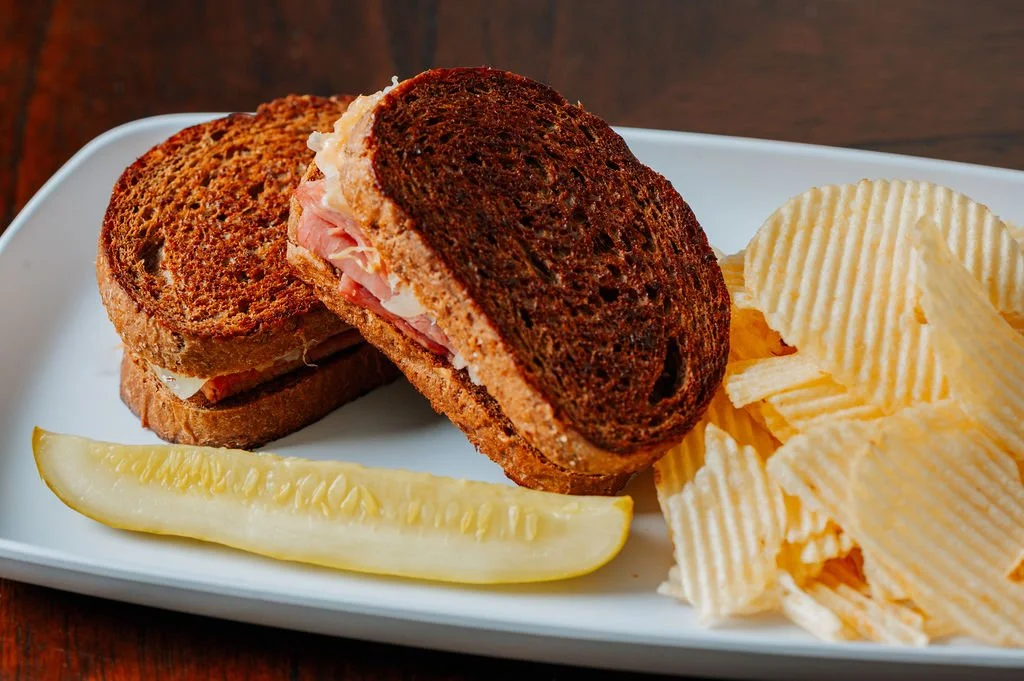 A plate with a toasted sandwich on rye bread cut in half, ridged potato chips, and a pickle spear.