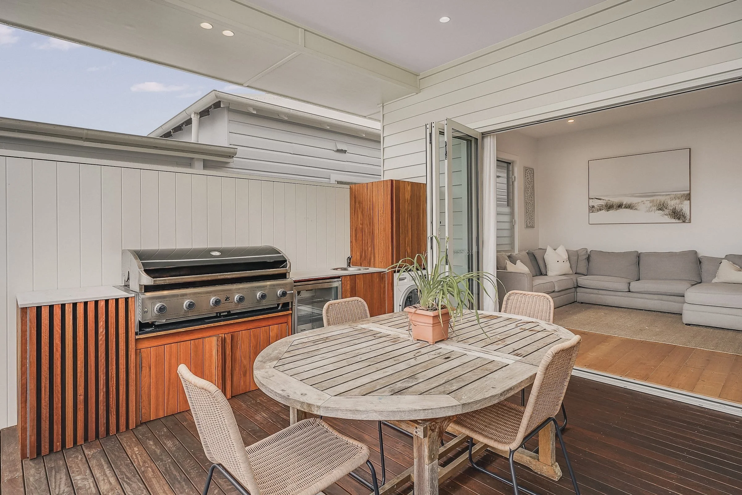 An outdoor patio area with a wooden dining table and four chairs, a built-in grill with wooden paneling, and a view of a living room with a gray sectional sofa and wall art through sliding glass doors.