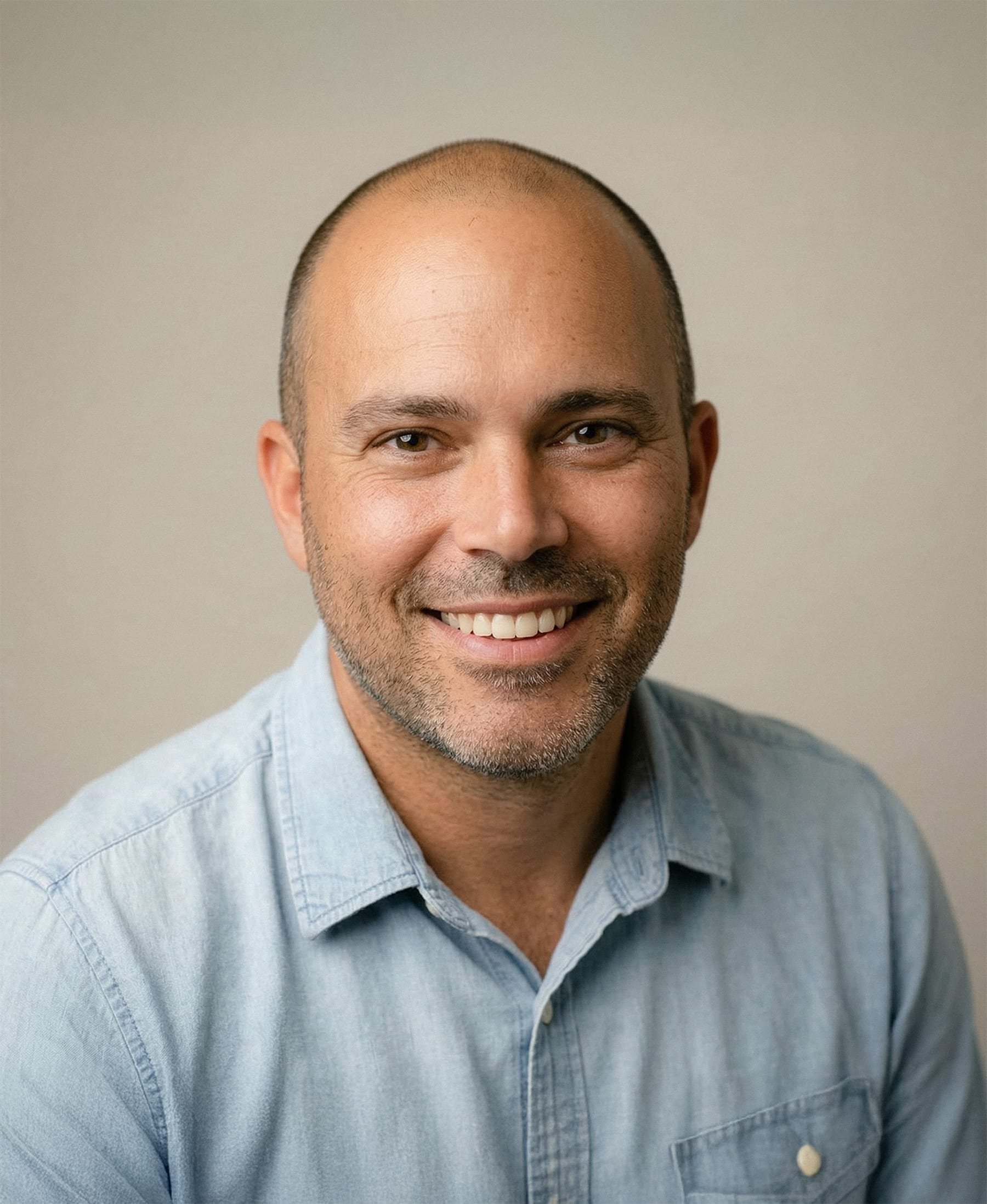 Close-up of a smiling man with a shaved head and brown eyes, wearing a light blue denim shirt, against a plain neutral background.