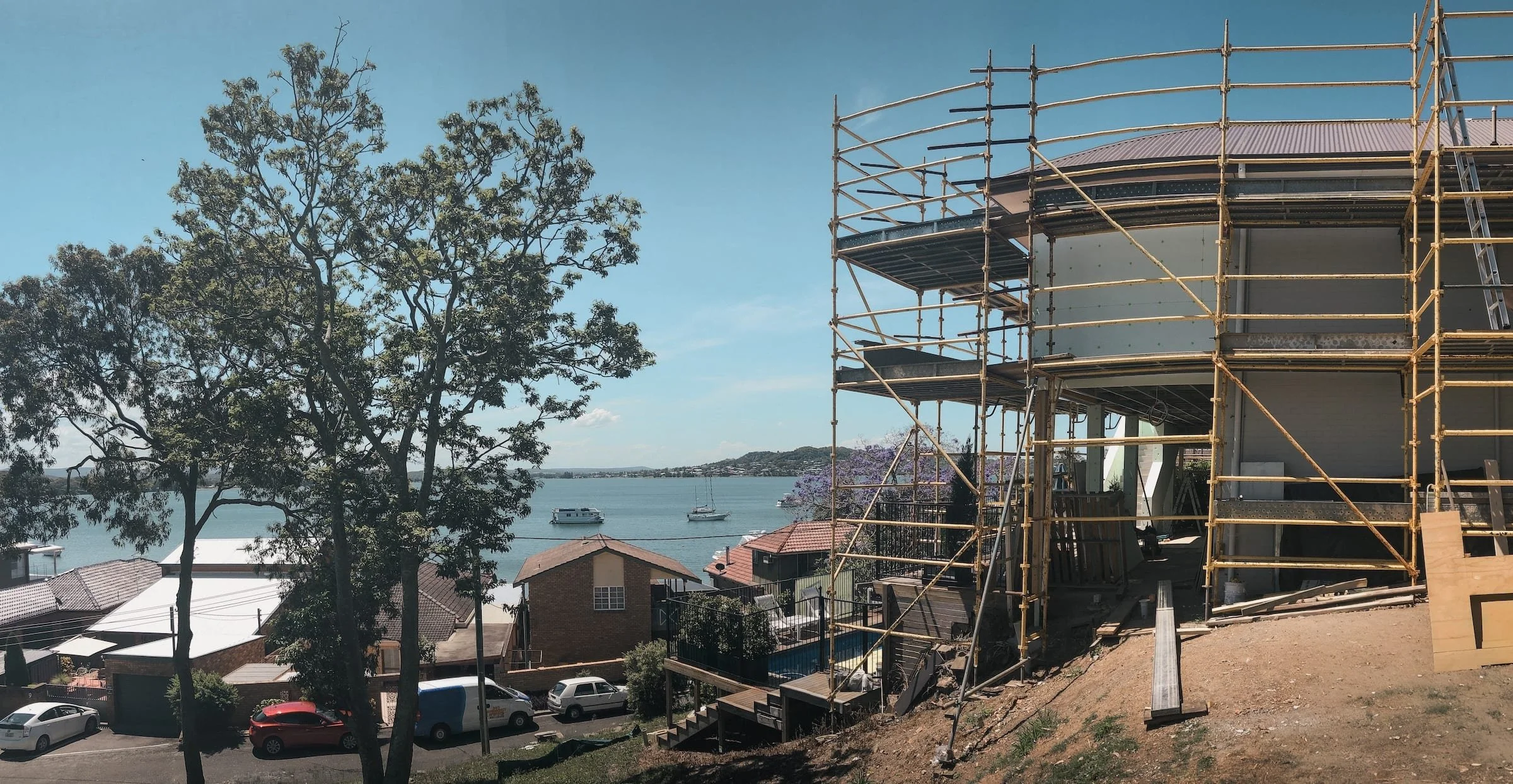 Construction scaffolding surrounding a house near water with boats, trees, and neighboring houses in a suburban area.