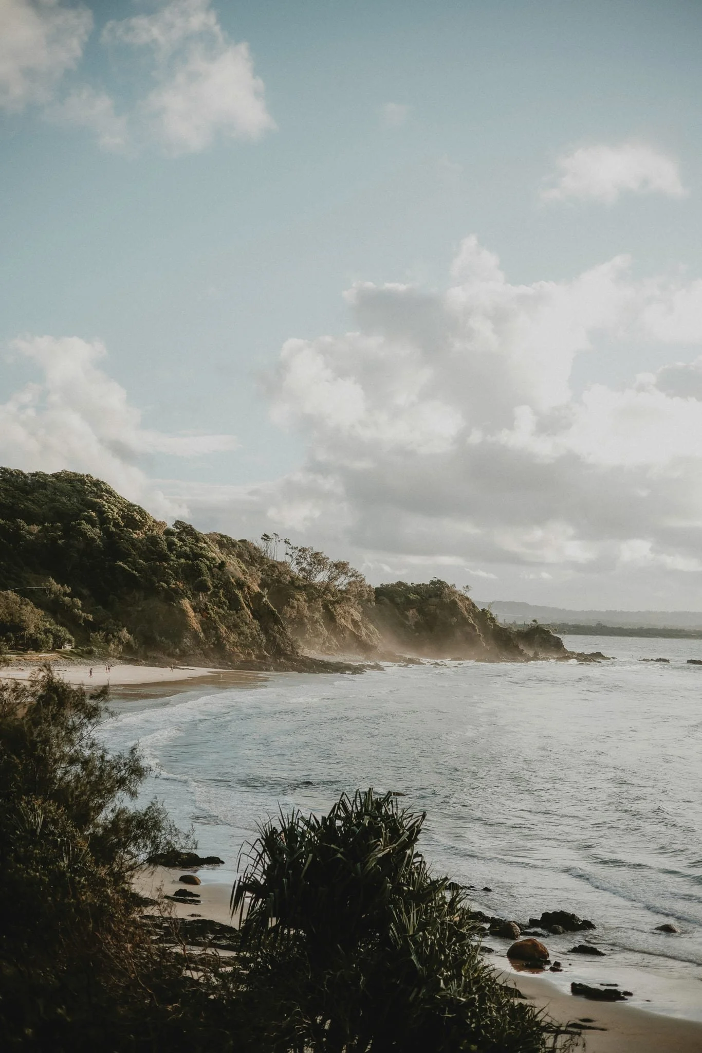 A scenic coastline with a sandy beach, rocky shoreline, lush green trees and bushes, and a cloudy sky over the ocean.