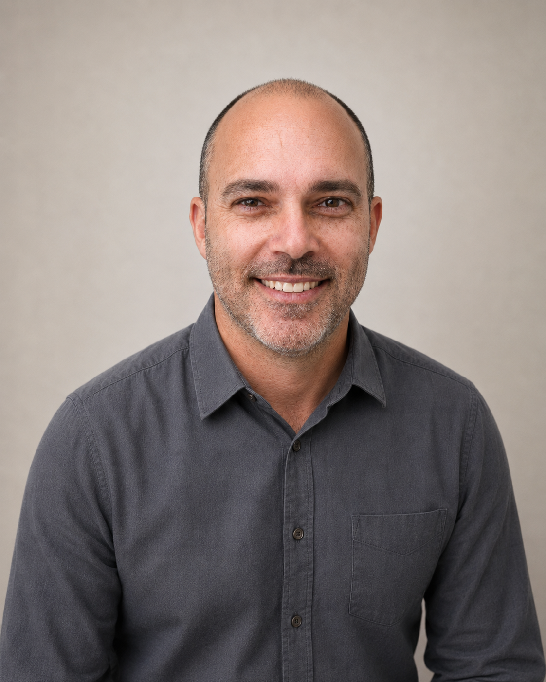 A smiling man with a shaved head, stubble, wearing a dark gray button-up shirt, against a plain light gray background.