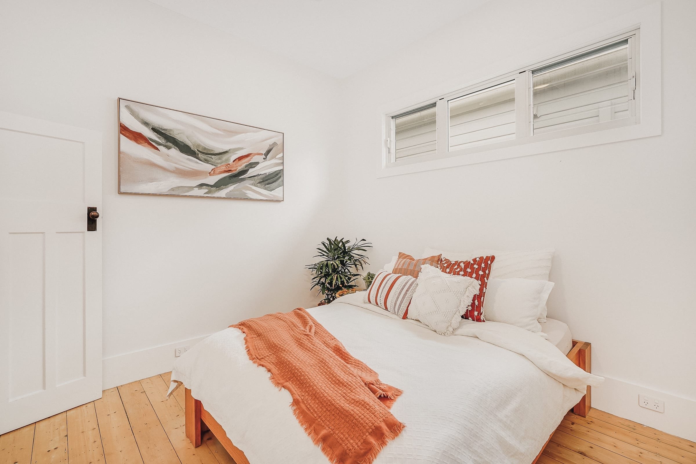 A cozy bedroom with a bed covered in white bedding and adorned with a variety of pillows in red and white patterns. An orange blanket is draped across the foot of the bed. Above the bed, there is an abstract painting with shades of white, gray, black, and red. To the right of the bed, a green houseplant is visible. The room features white walls, a wooden floor, a small window with horizontal blinds, and a closed white door on the left side of the image.