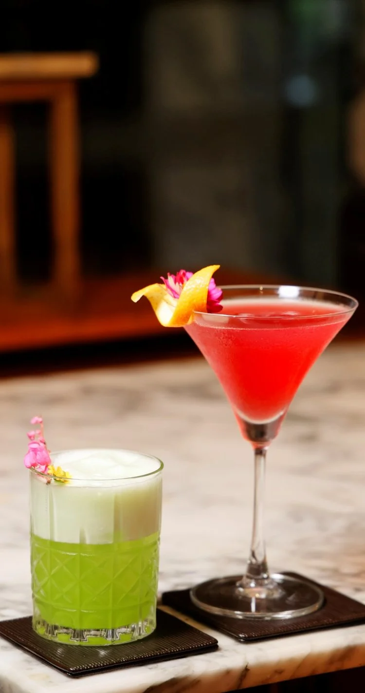 Sophisticated mixology presentation featuring vibrant red martini and green sour cocktail on marble surface.