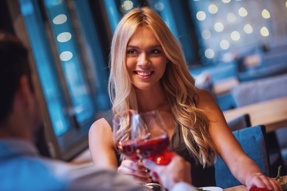 Beautiful young woman is looking at her guy friend, drinking wine and smiling while they having a date in restaurant