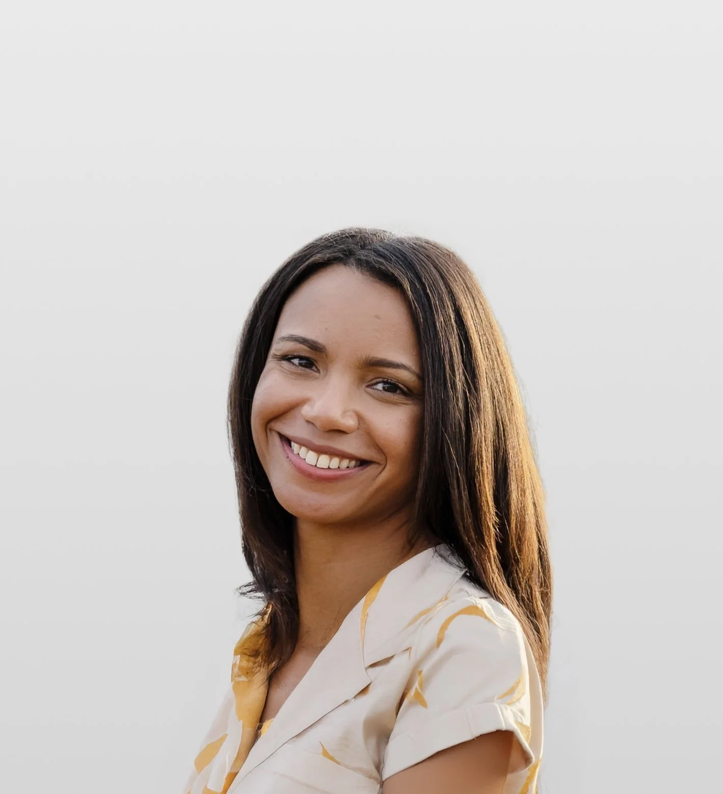 A smiling woman with dark brown hair and wearing a cream-colored shirt with yellow patterns, stands against a plain light gray background.
