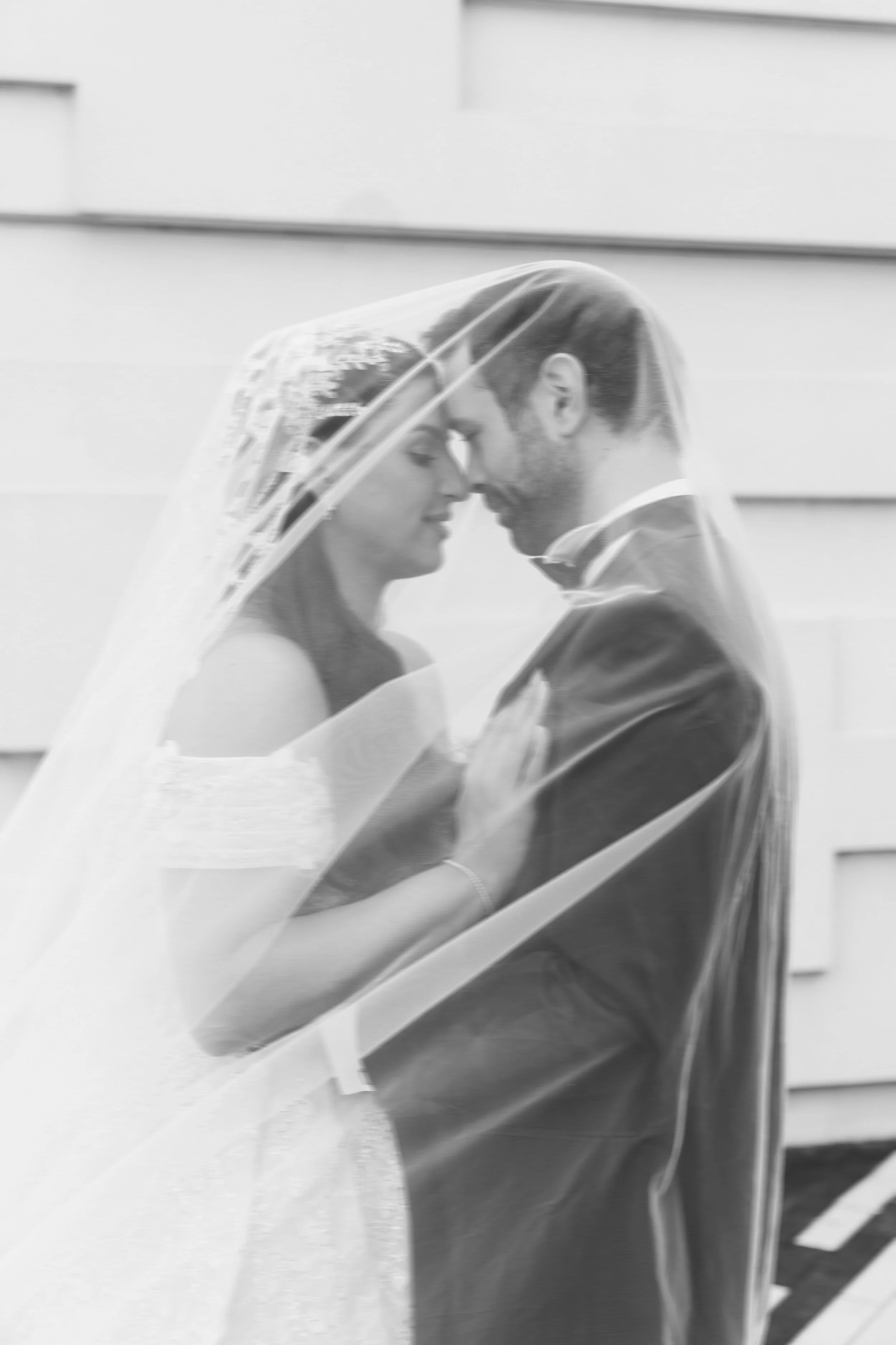 A black-and-white photo of a couple embracing and touching foreheads with their eyes closed, partially covered by a wedding veil.