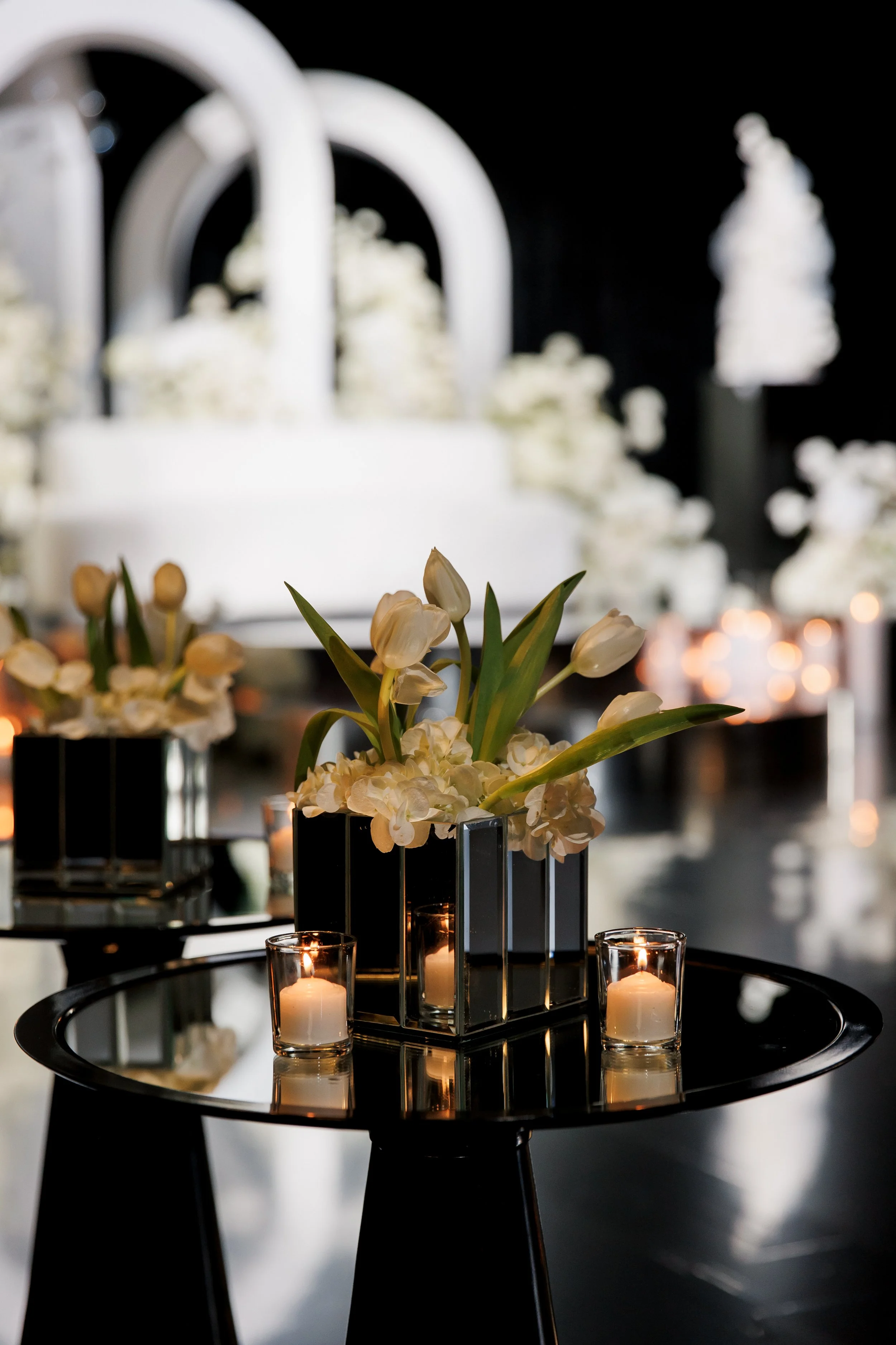 A floral centerpiece with white tulips and white roses inside a black geometric vase, surrounded by candles on a reflective black table, with blurred floral arrangements in the background.