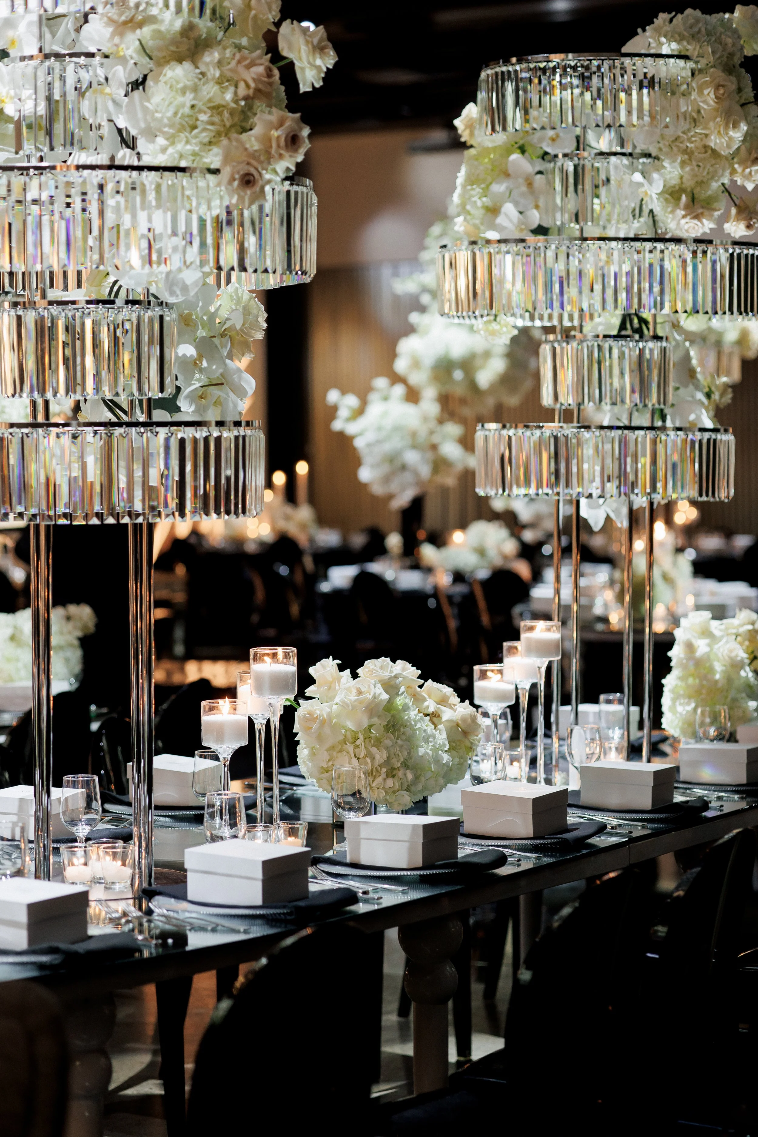 Elegant banquet table setup with white floral centerpieces, candles, and crystal chandeliers in the background, suggesting a wedding or formal event. Large Crystal Chandeleir centrepieces. Custom mirror oval Tables. Custom design.