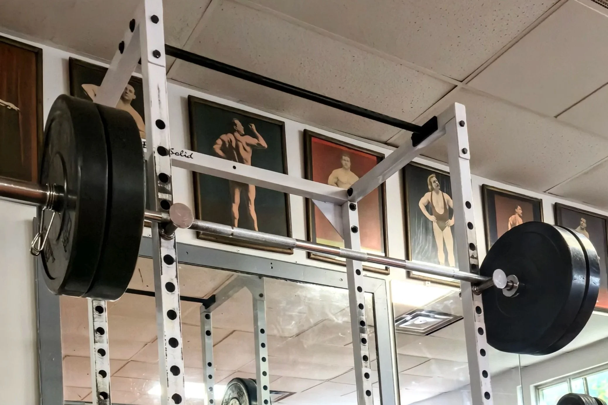 A power rack with a barbell loaded with weight plates in a gym. Framed bodybuilding photos on the wall in the background.