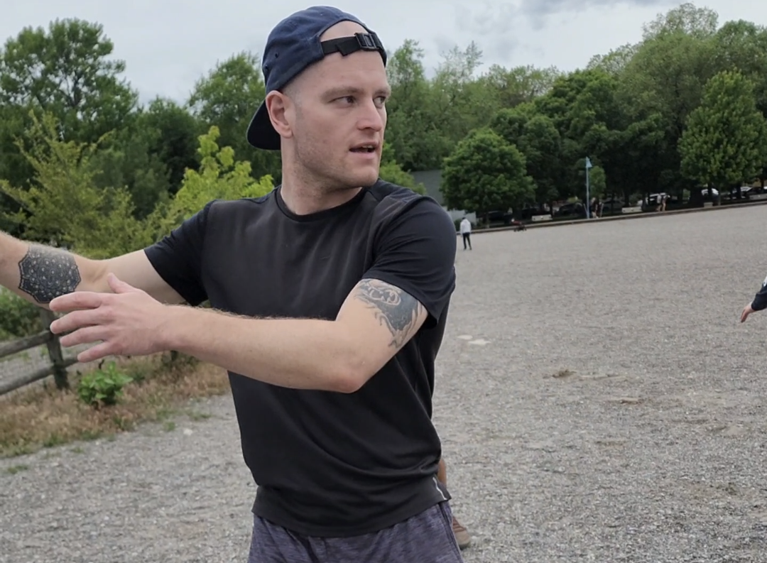 A young man with a baseball cap and tattoos on his arms, wearing a black t-shirt, standing outdoors on a gravel area, gesturing with his arms, with trees and a few people in the background.