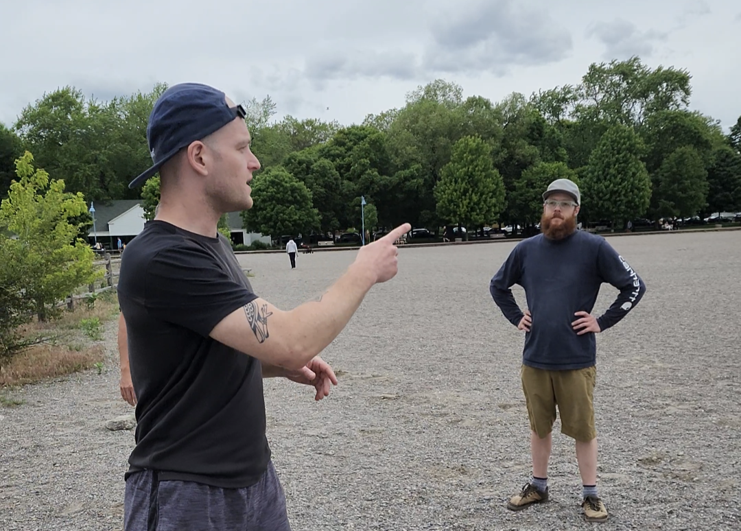 Two men standing outdoors on a gravel area, one man is pointing and speaking to the other, with trees and a few people in the background.