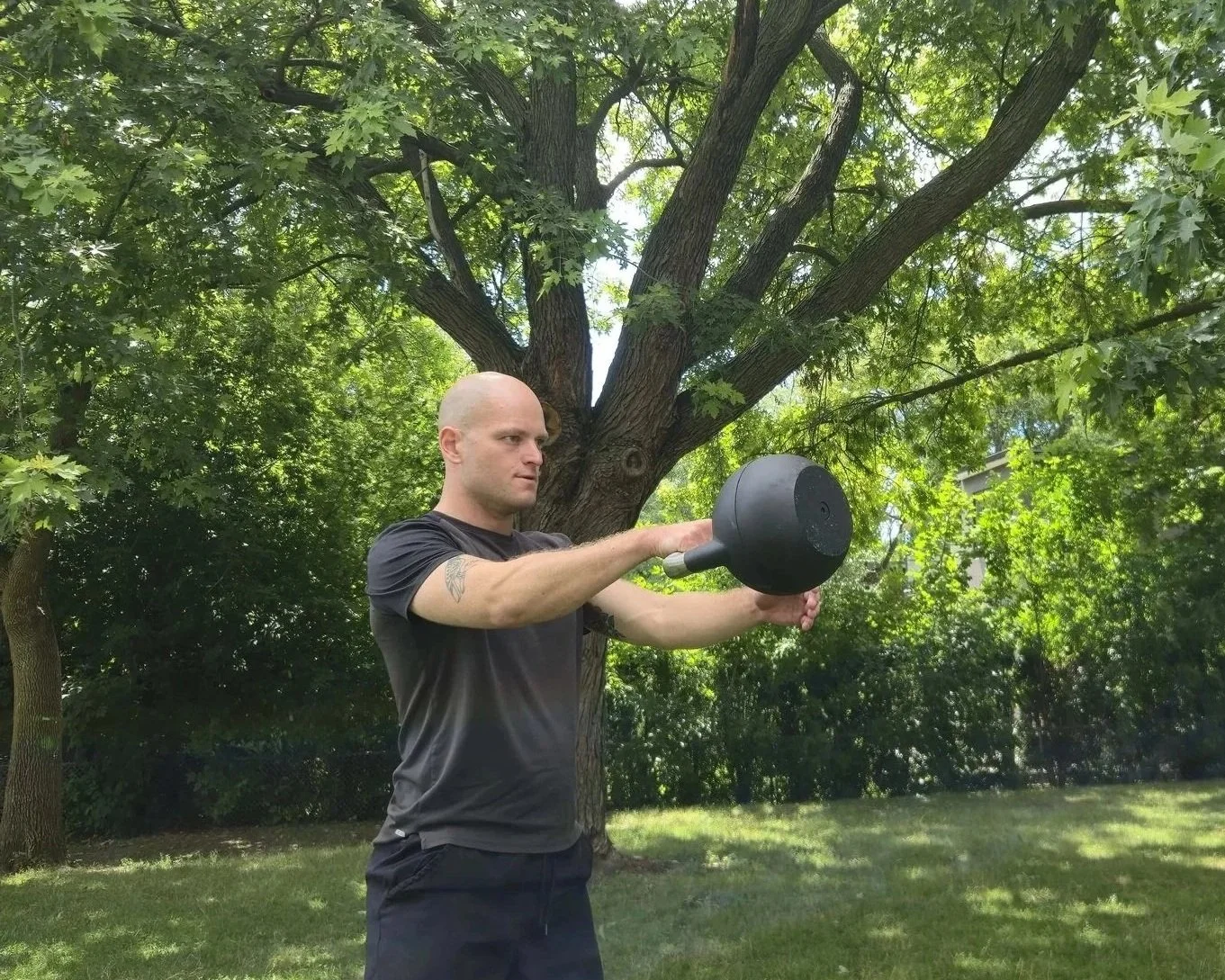 A man with a bald head and tattoo on his right arm lifting a dumbbell in a park with green trees and grass.