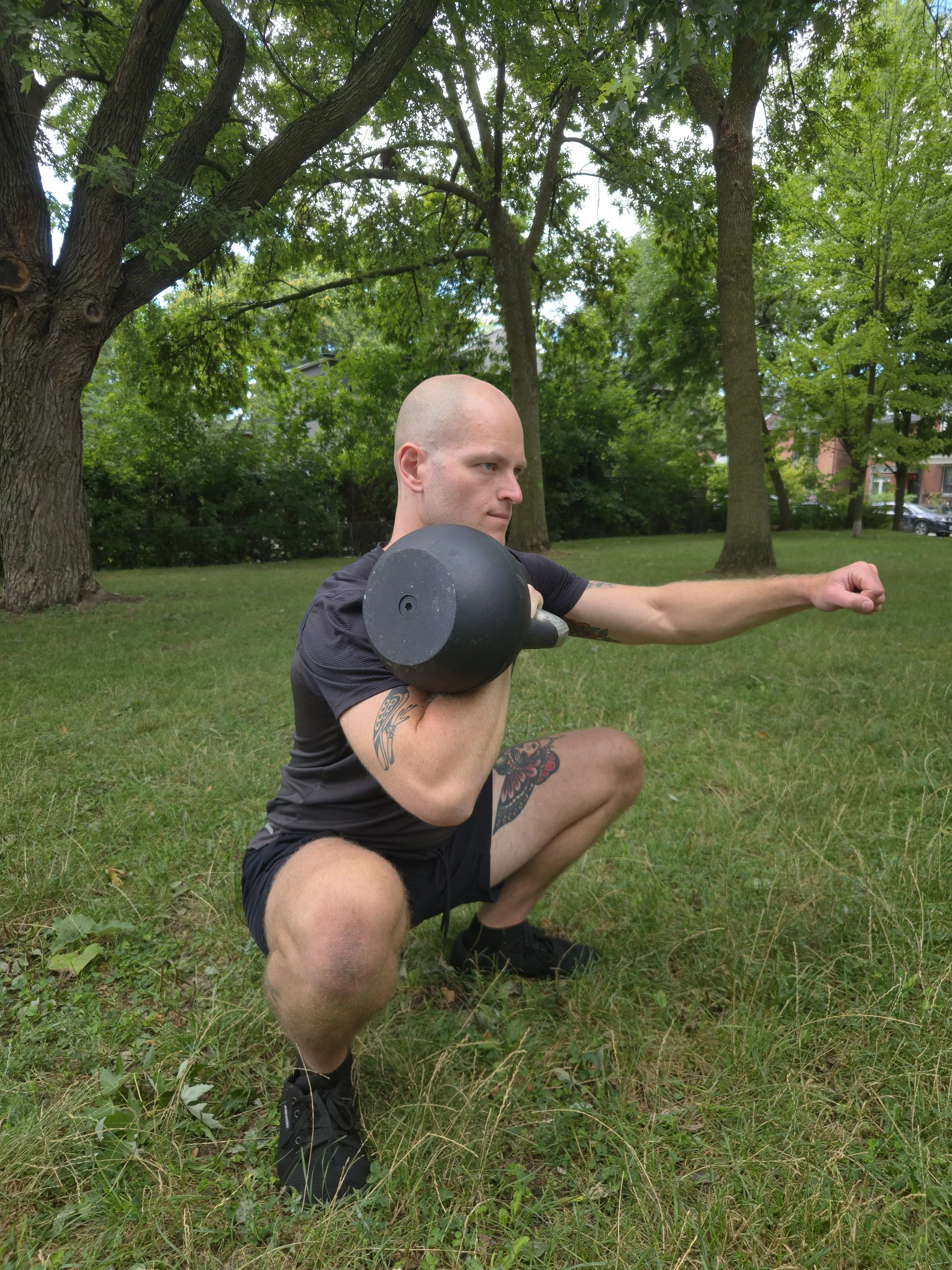 A man with tattoos squatting outdoors on grass, holding a kettlebell on his shoulder, extending his other arm forward.