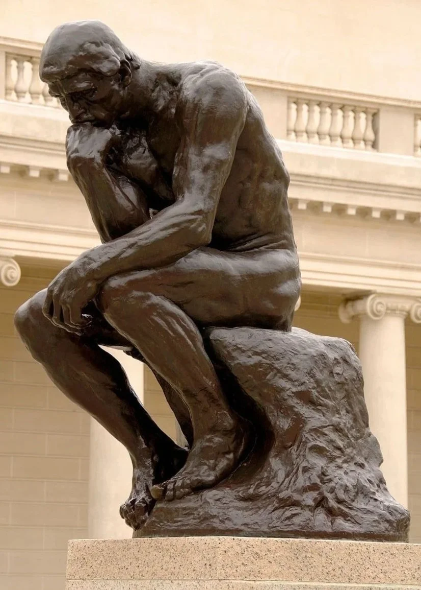 A bronze sculpture of a man sitting on a rock with his chin resting on his hand, front view, in a museum setting with classical architecture.