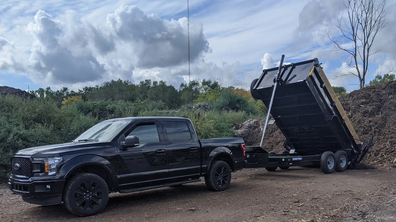 Black pickup truck attached to a dump trailer on dirt ground, with the trailer bed raised.