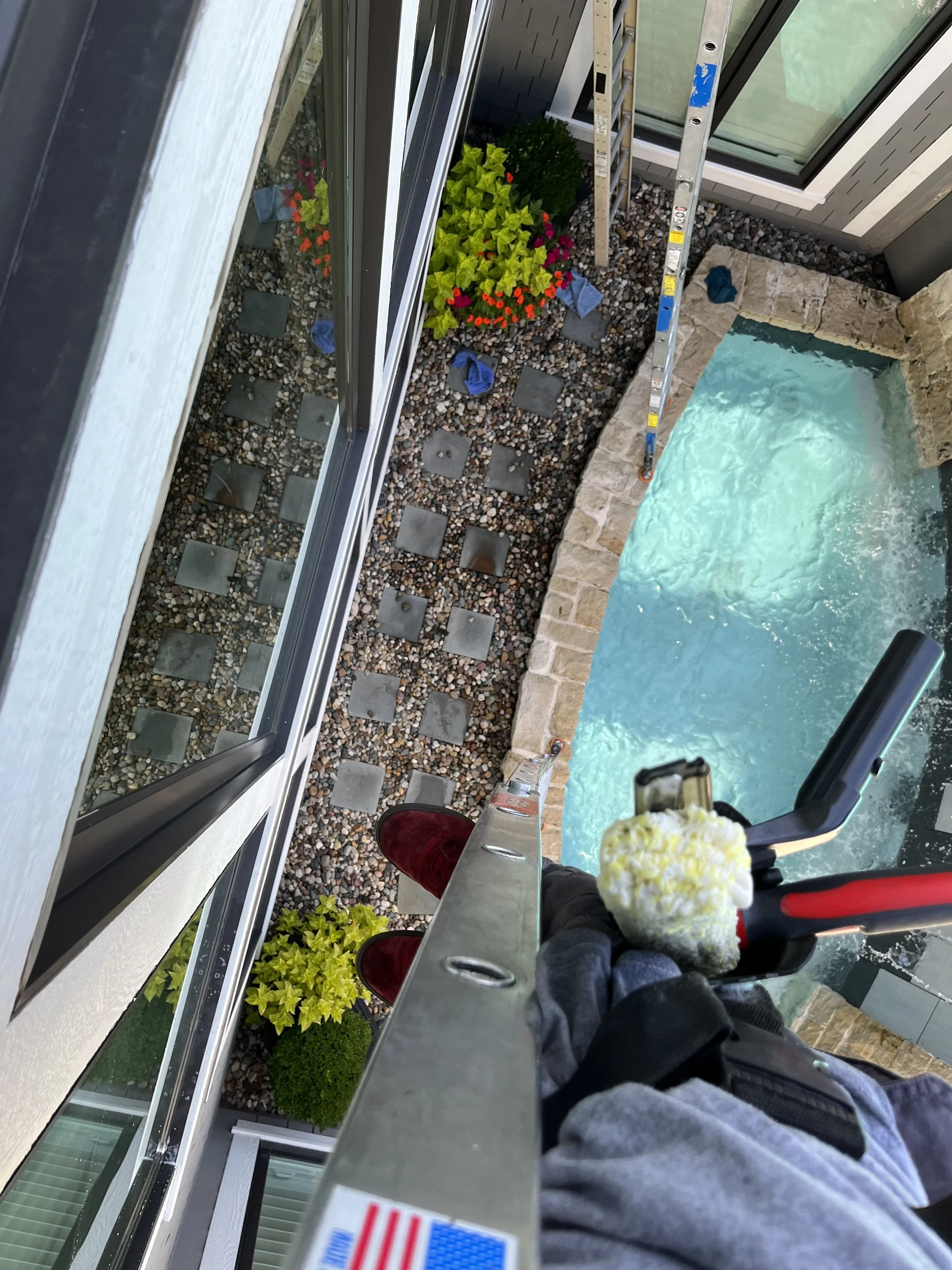 Person working on window installation or cleaning from a ladder, with a ladder, a pool, and landscaping below.