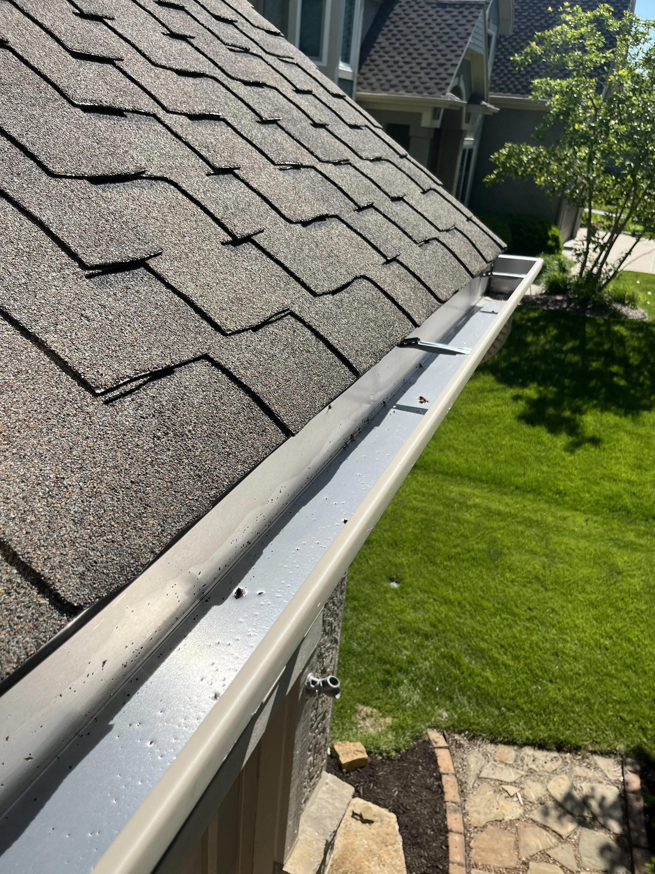 Close-up view of a house roof with brown asphalt shingles and a light-colored metal gutter system, with a well-maintained green lawn and some trees in the background.