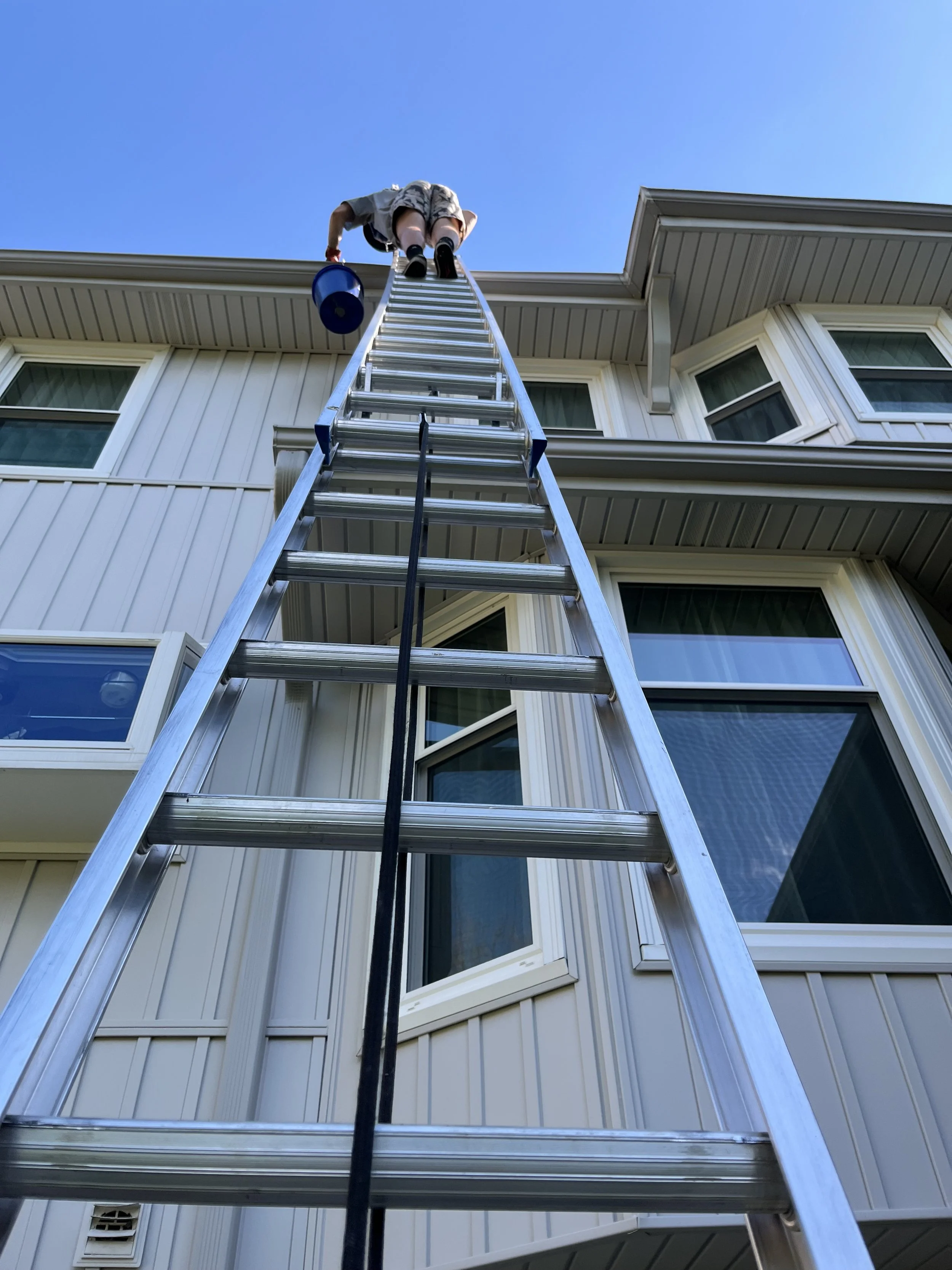 Person on a tall extension ladder cleaning or inspecting the exterior of a multi-story house with beige siding and white-framed windows under a clear blue sky.