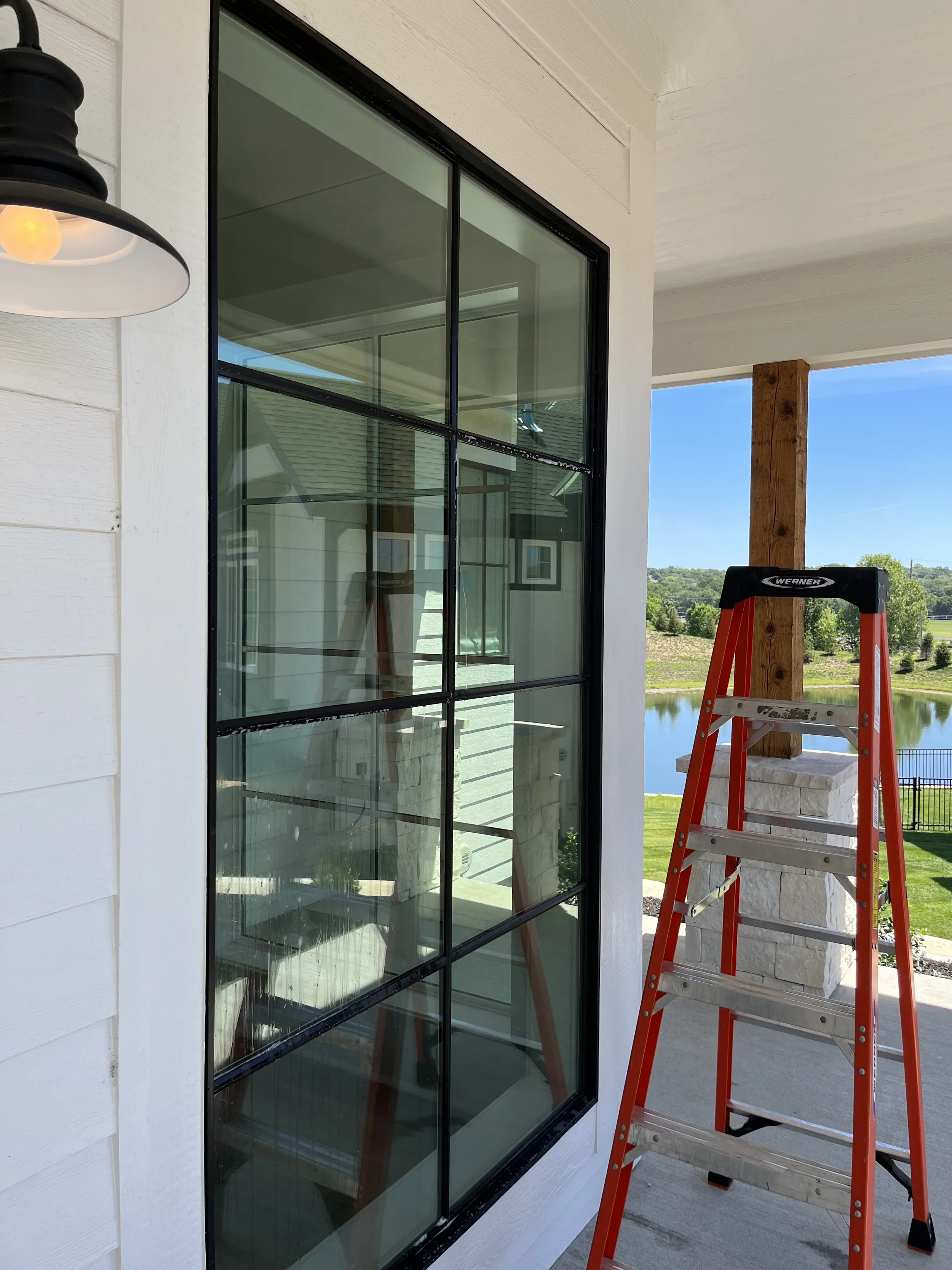 A large window with black metal grid panes on a white house exterior. A red step ladder is positioned nearby on the porch, overlooking a scenic view of a lake, greenery, and clear blue sky.