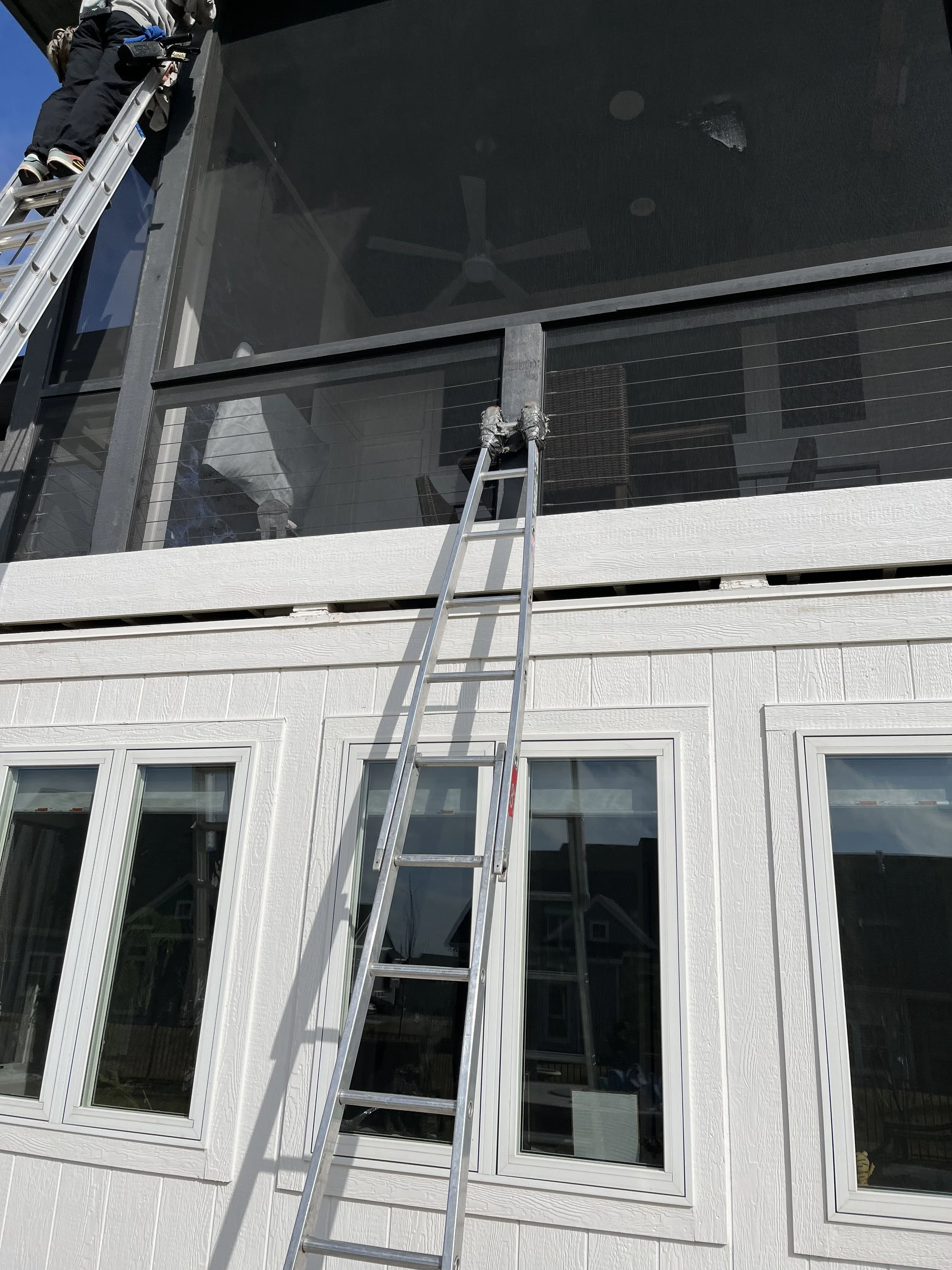 A ladder leaning against the exterior of a house, with a worker at the top on a balcony or deck, working on the house's upper part.