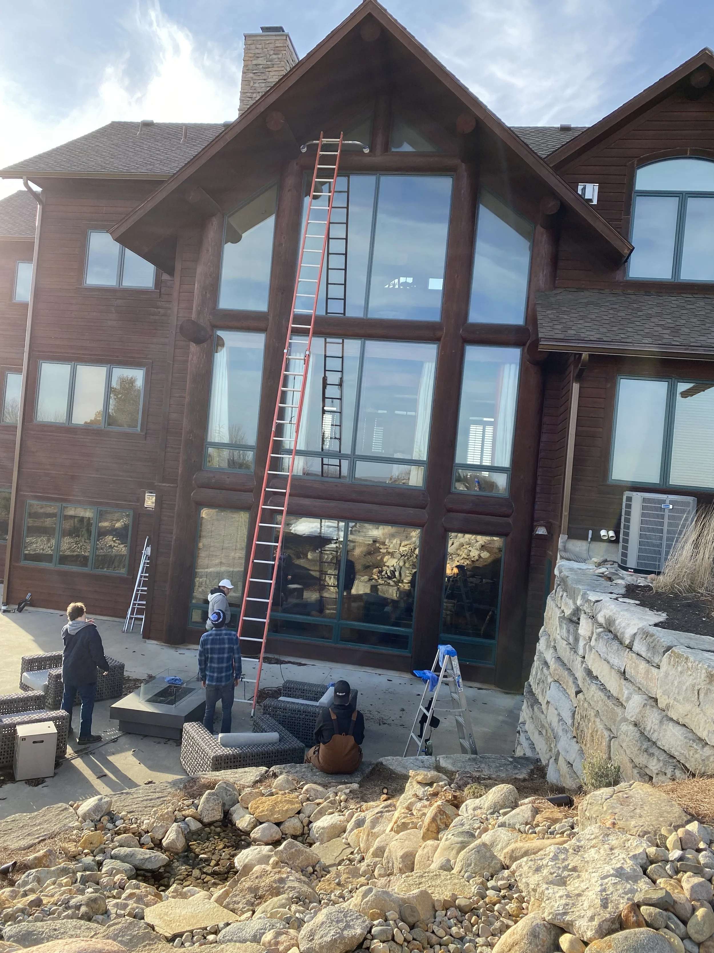 Construction workers installing a window in a modern multi-story wooden house with large glass panes and a steeply pitched roof, with outdoor seating and stone landscaping in the foreground.