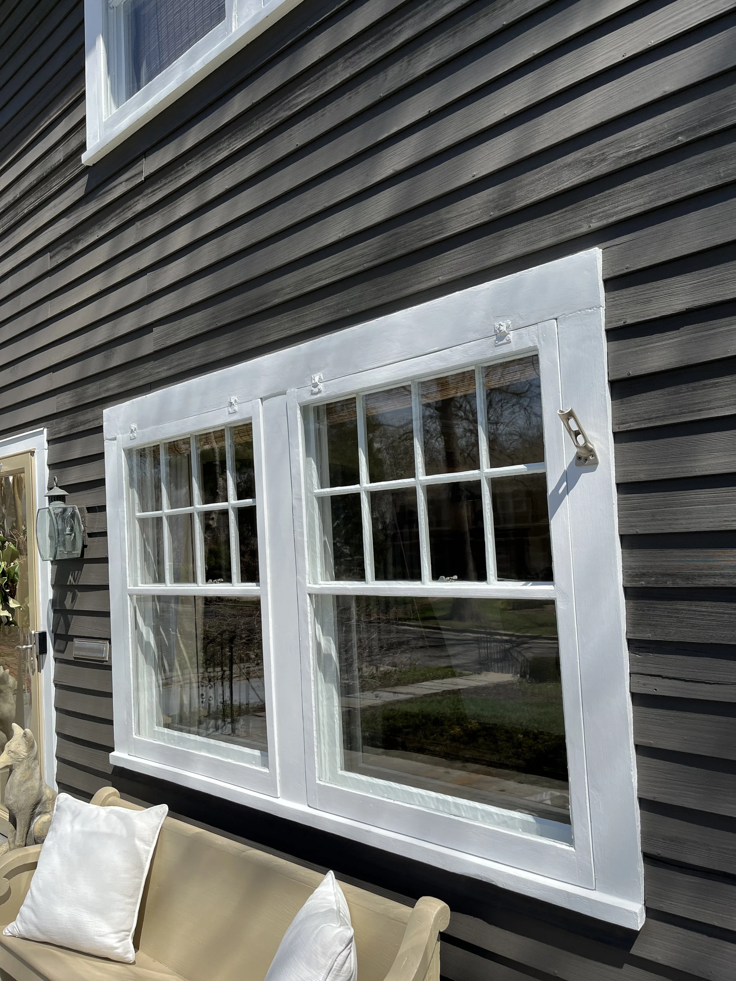 Close-up of a house exterior featuring a black wooden siding wall, a white-framed double-pane window, and outdoor furniture with white cushions.