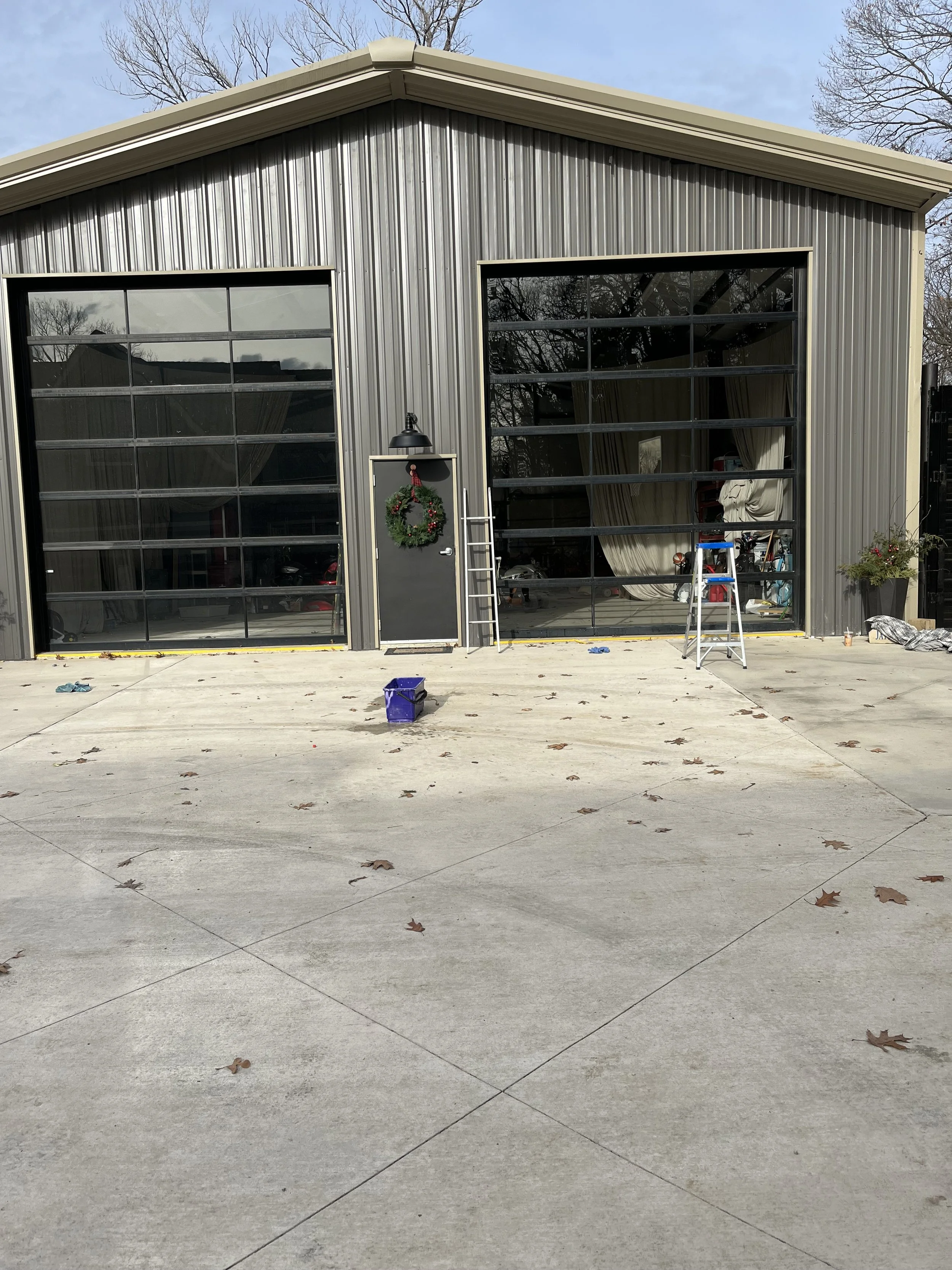 Industrial-style metal garage with large glass doors, one decorated with a Christmas wreath. The driveway in front is concrete with scattered fallen leaves. There are two ladders and a purple container on the driveway, with tools and supplies visible inside through the glass doors.