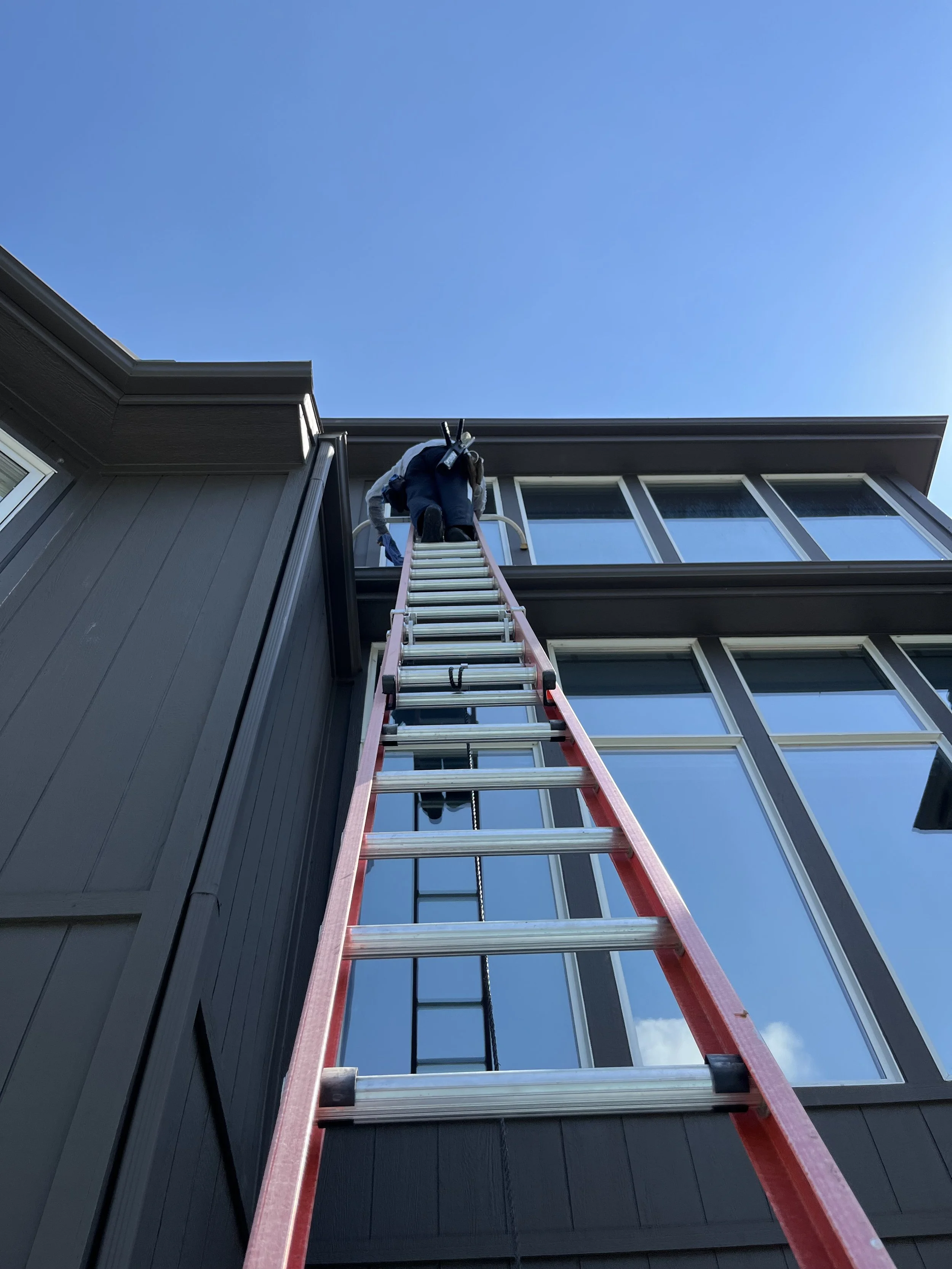 A person is climbing a tall red extension ladder leaning against a dark gray house with large windows, reaching up to the roof under a clear blue sky.