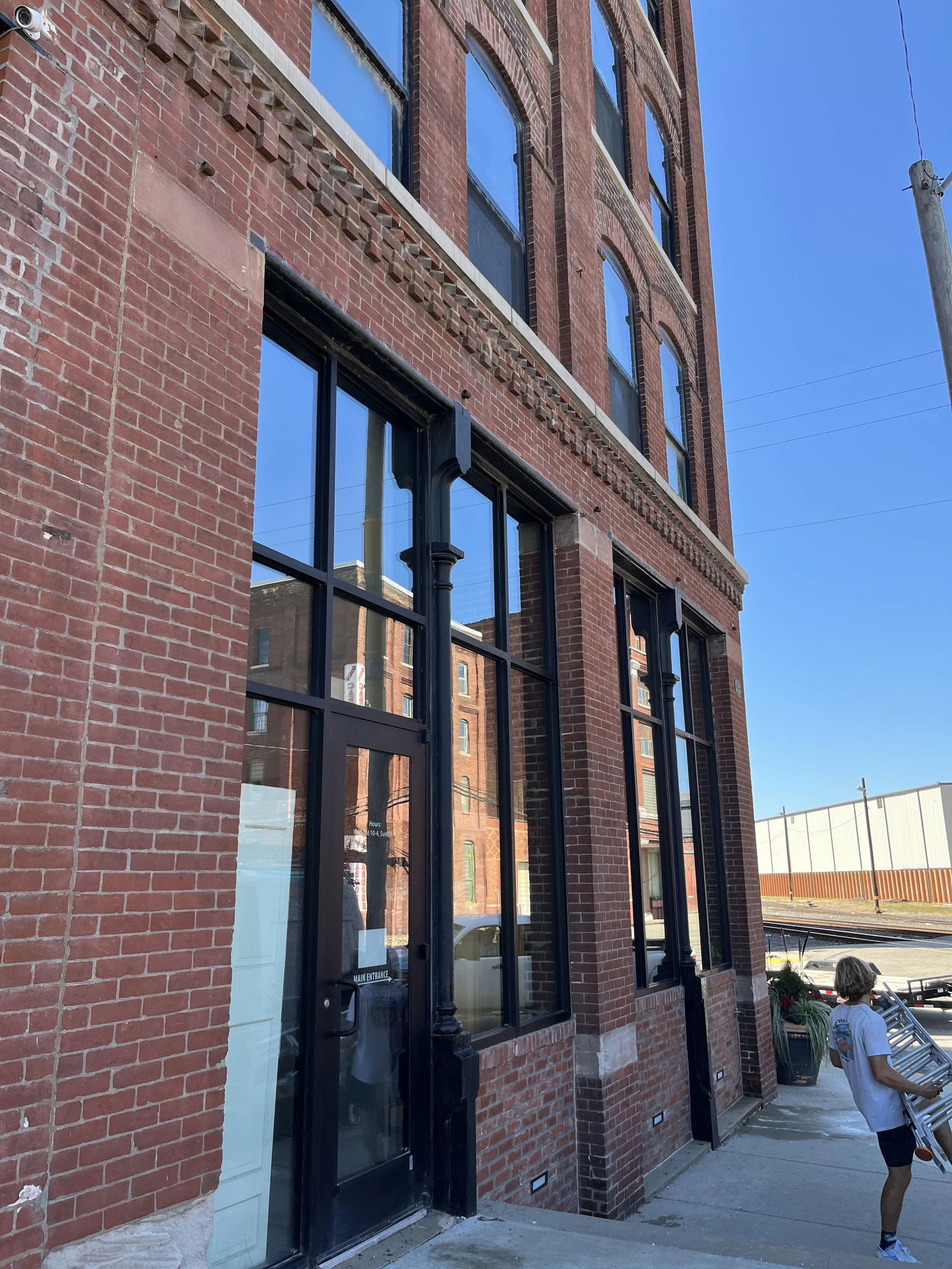Red brick building with large black-framed windows, a glass door labeled 'Main Entrance', and a person walking past carrying a metal object outdoors on a sunny day.