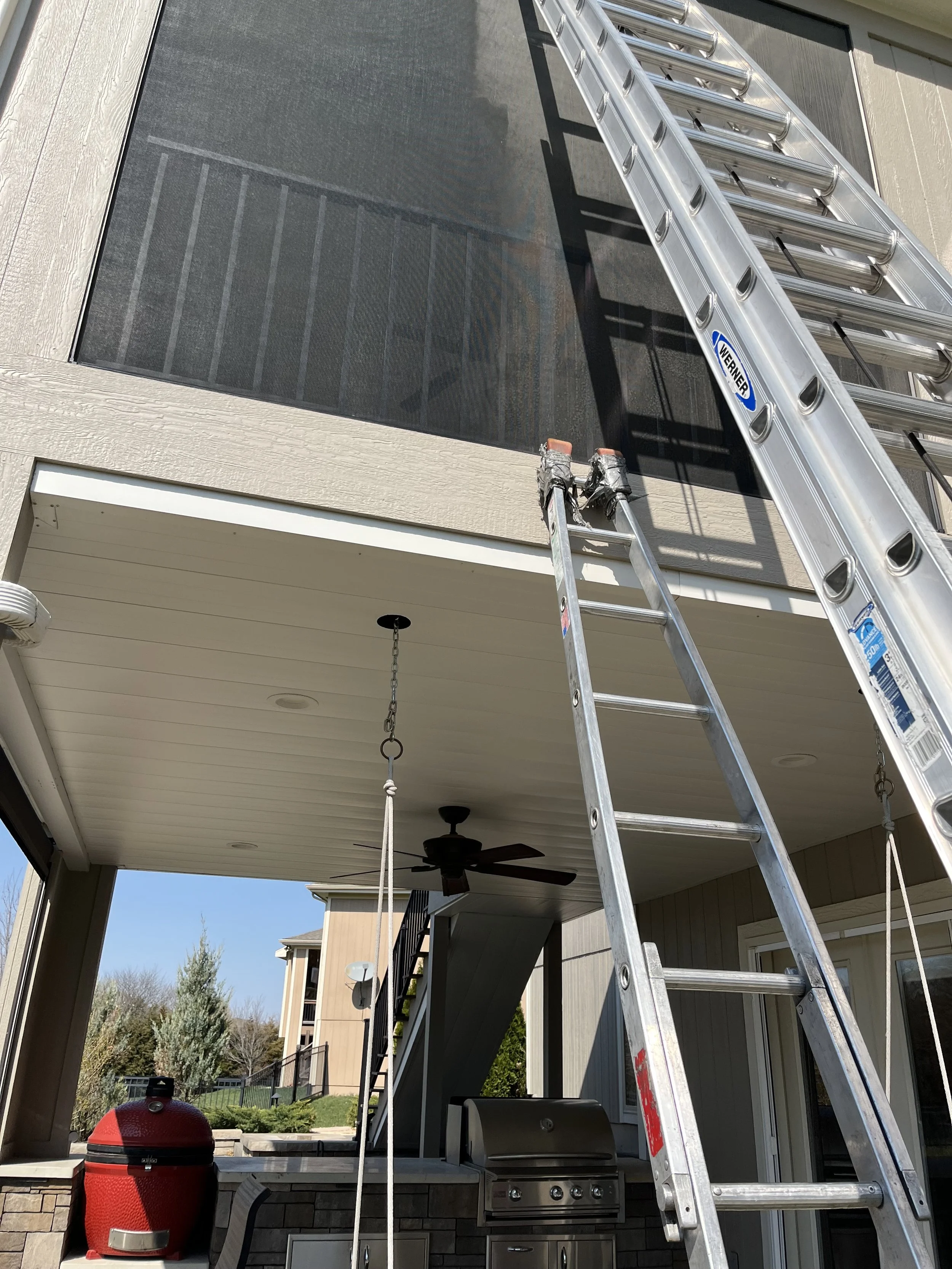 Ladder leaning against a screened porch outside house, with another extension ladder on top of it, and outdoor ceiling fan, grill, and patio area visible underneath.