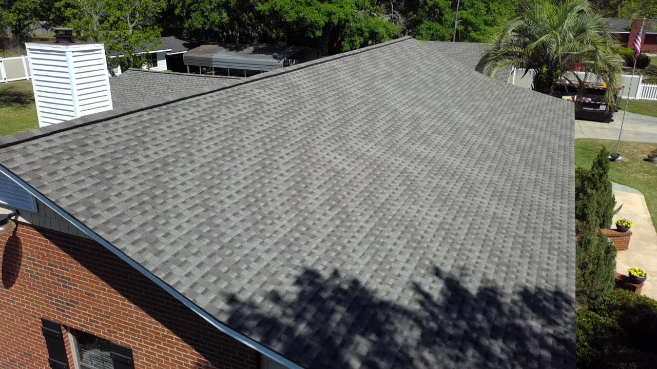 Aerial view of a house's grey shingle roof, with surrounding trees, a brick exterior wall, and a front yard with potted plants.